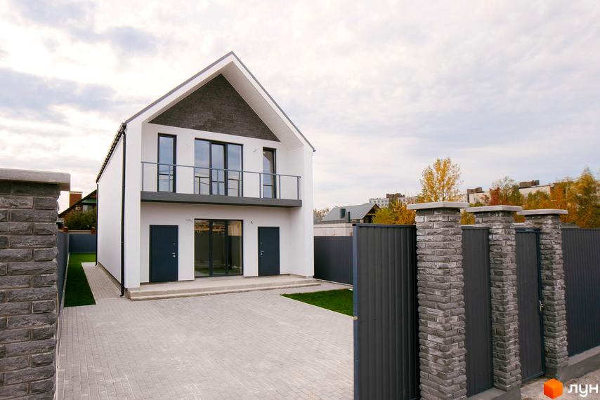 Білий двоповерховий будинок Котеджного містечка Щастя з темним дахом та балконом. Територія має бруківку, газон та паркан з цегляними стовпами.