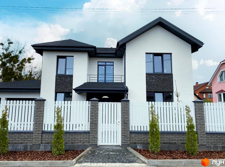Білий двоповерховий будинок Котеджного містечка Щастя з темним дахом та цегляними вставками. Перед будинком білий паркан з кам'яними стовпами та зелені насадження.