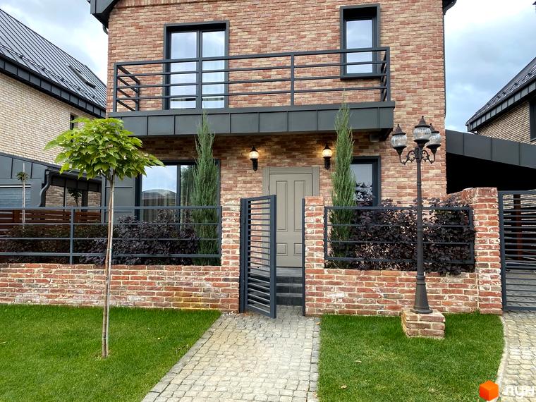 Цегляний будинок Котеджного містечка Щастя з темними вікнами та балконом. Навколо будинку зелений газон, бруківка, цегляний паркан та вуличний ліхтар.