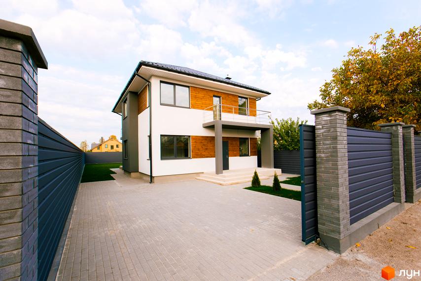 Двоповерховий будинок з дерев'яним оздобленням на фасаді. Перед будинком викладена бруківка, є газон та огорожа з блоків.