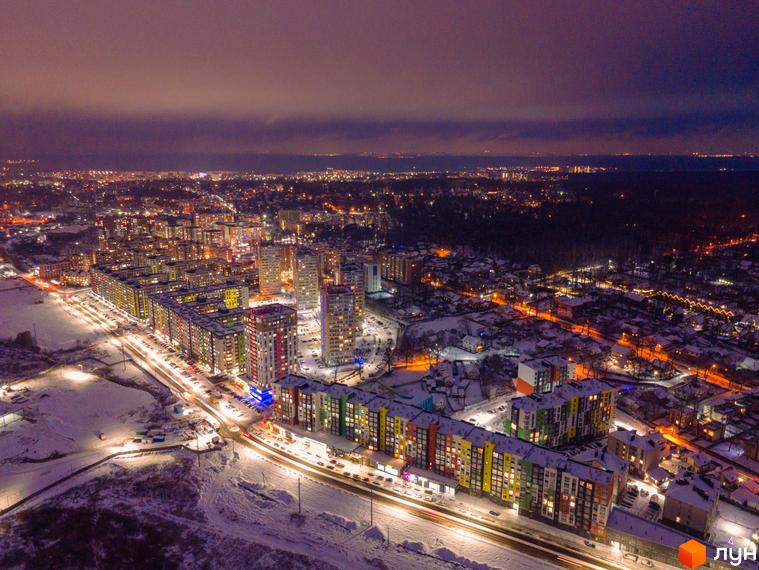 Нічний вид на ЖК Синергія-3+ з яскравими фасадами будинків та засніженою територією. Комплекс розташований біля освітленої дороги з автомобілями.