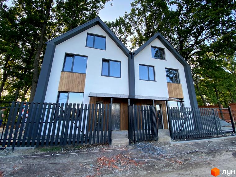 Двоповерховий дуплекс з білими стінами, дерев'яними вставками та темним дахом. Перед будинком чорний паркан та територія з будівельними матеріалами.