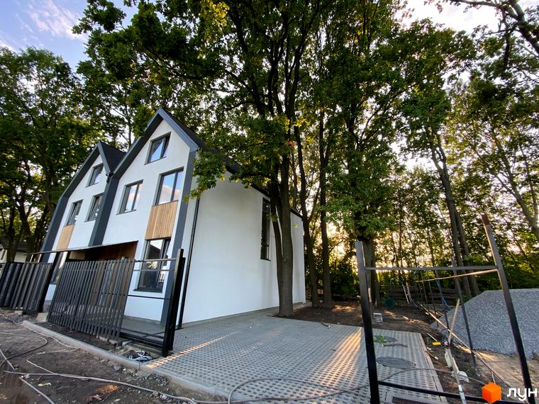 Білосніжний дуплекс з темним дахом на тлі зелених дерев. На території ЖК Дубки облаштовано парковку та встановлено огорожу.