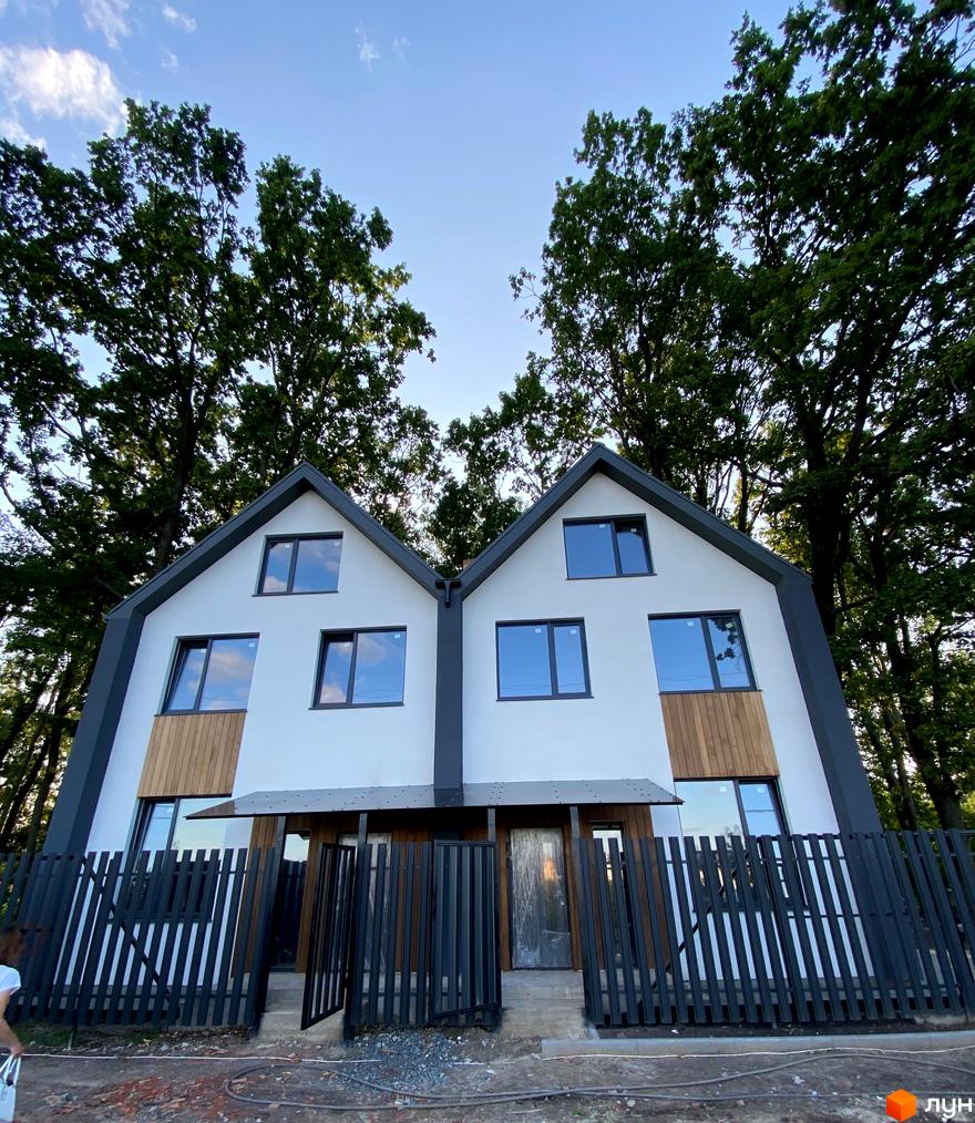 Двоповерховий будинок з білими стінами, темною покрівлею та дерев'яними вставками на фасаді. Перед будинком розташований чорний паркан та невелика прибудинкова територія.