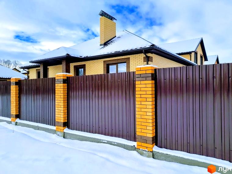 Котеджне містечко Green city: будинок з жовтої цегли, темним дахом та вікнами. Перед будинком коричневий паркан з цегляними стовпами, вкритий снігом.