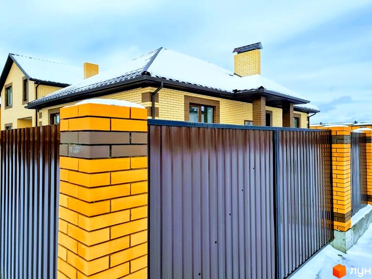 Будинок в Котеджному містечку Green city з світло-жовтої цегли та коричневим дахом. Прибудинкова територія має паркан з профнастилу та цегляних стовпів.