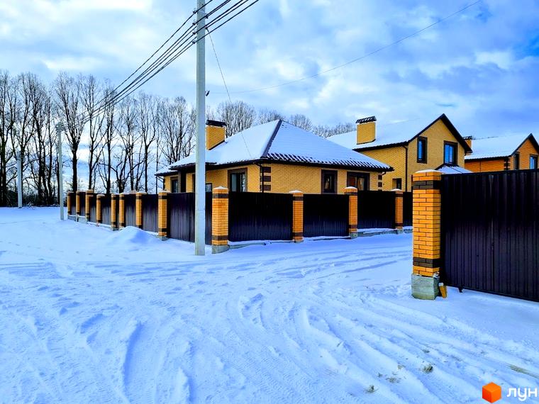Котеджне містечко Green city: будинки з жовтої цегли з темними дахами, вкритими снігом. Прибудинкова територія зі снігом, парканом з цегляними стовпами та деревами на фоні.