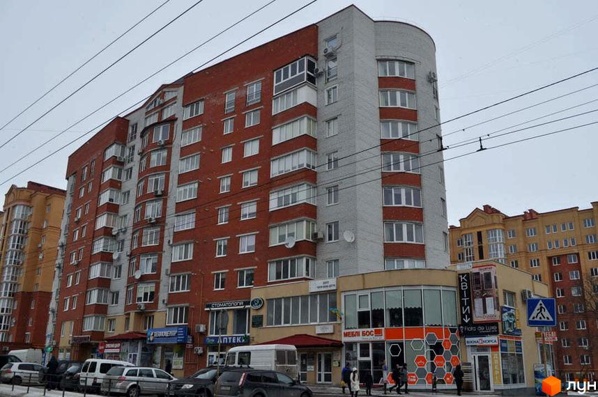 Житловий комплекс на вул. Київська, 7д – багатоповерховий будинок з червоно-сірим фасадом, магазинами на першому поверсі та вулицею з авто.
