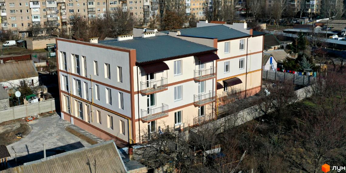 Триповерховий будинок з балконами та темно-зеленим дахом. Поруч розташовані дерева та інші будівлі.