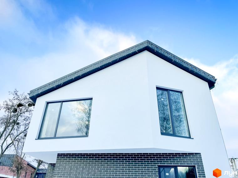 Зовнішній вигляд білого будинку з темною цегляною основою та великими вікнами. Навколо будинку видно дерева та блакитне небо з хмарами.