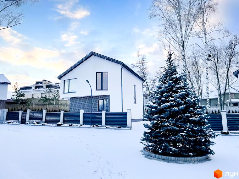 Білий будинок з темними вікнами та дахом на тлі засніженого двору. Поряд з будинком росте ялинка, а територія огороджена парканом.