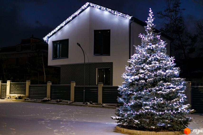 Будинок з білими стінами та гірляндою на даху, перед будинком стоїть ялинка, територія вкрита снігом. Котеджне містечко Friends Club.