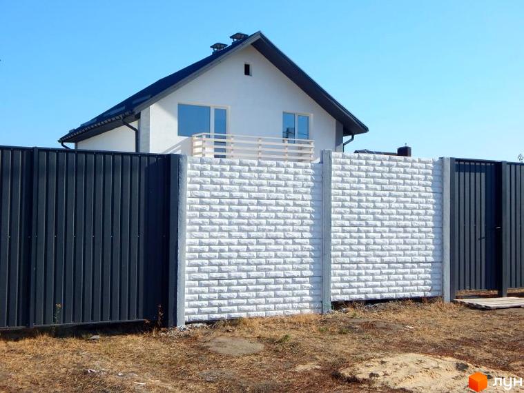 Зображення будинку з білими стінами та чорним дахом, огородженого парканом. На території видно суху траву та блакитне небо.