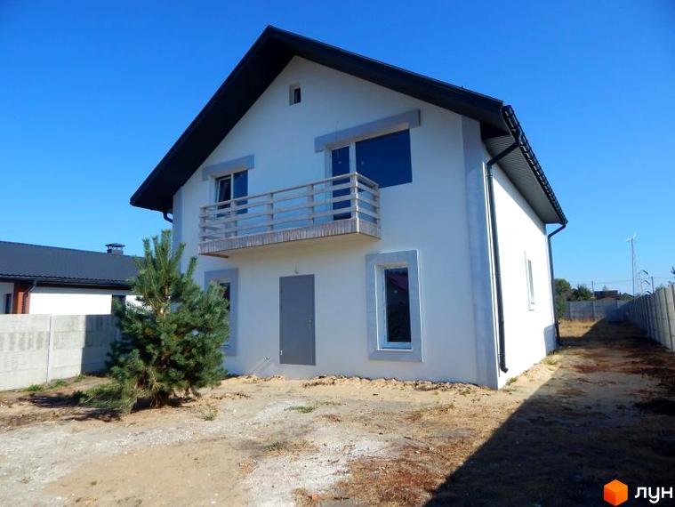 Двоповерховий будинок з білими стінами, чорним дахом та балконом. Поруч з будинком росте ялинка, а територія покрита піском.