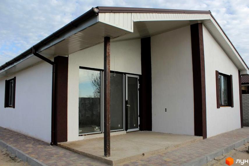 Білий будинок з коричневими акцентами та скляними дверима. Перед будинком бетонний майданчик, викладений тротуарною плиткою.