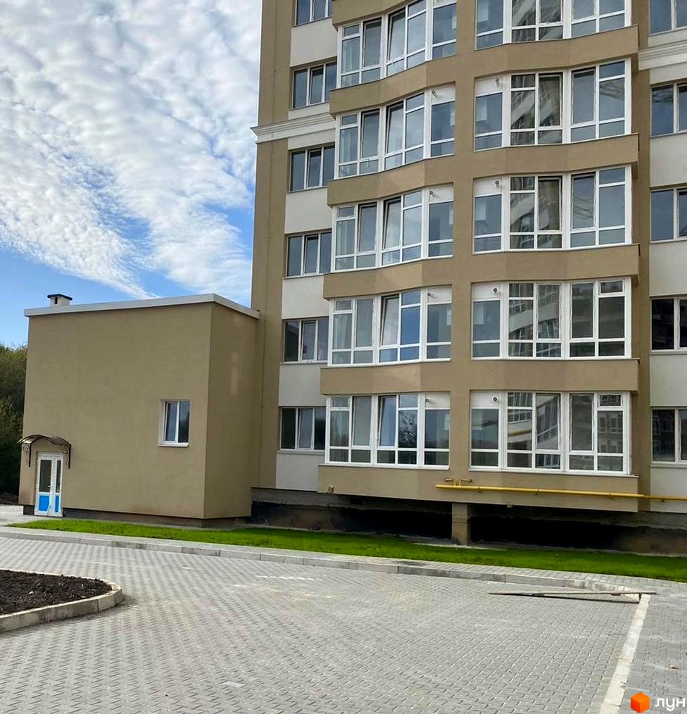 Зовнішній вигляд новобудови на вул. Зарічанська, 3/3б, з сірою фасадною обробкою та великою кількістю вікон. Прибудинкова територія облаштована бруківкою та зеленими насадженнями.