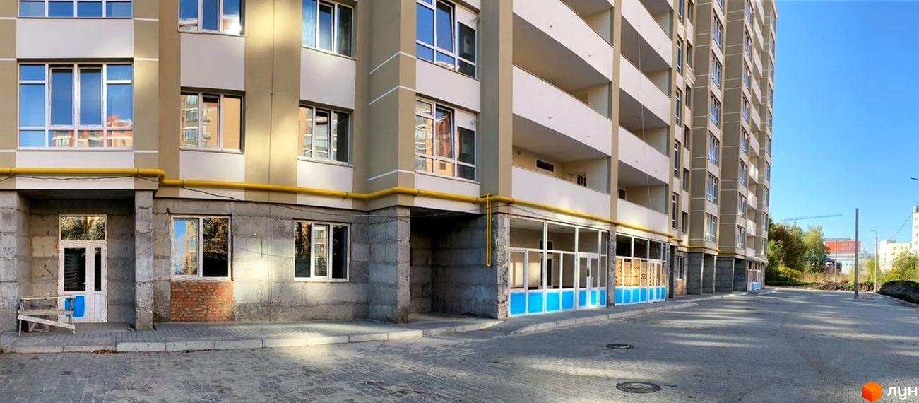 Багатоповерховий житловий будинок з балконами та вікнами, перший поверх з комерційними вікнами. Територія перед будинком вимощена бруківкою.