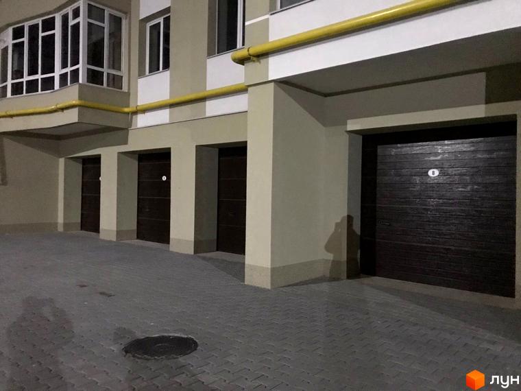 Фасад житлового будинку світлих тонів з жовтою трубою, балконом та темними гаражними воротами. Прибудинкова територія вимощена бруківкою.