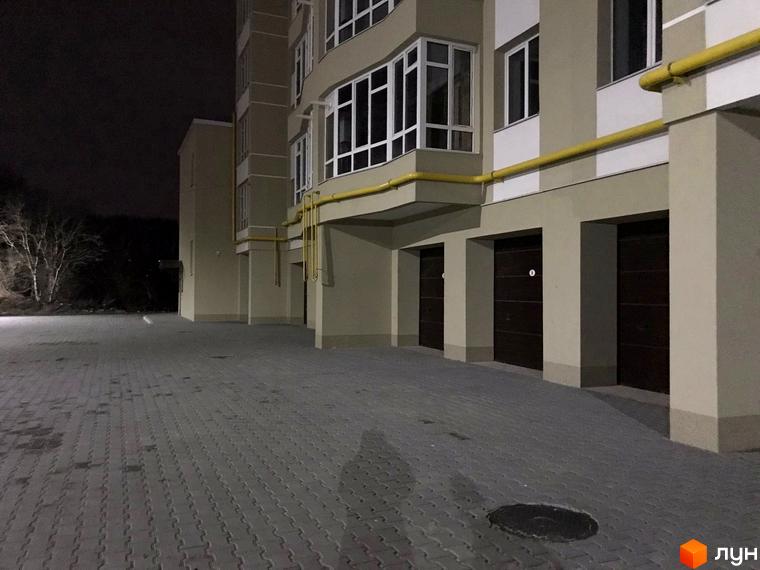 Нічний вигляд житлового комплексу на вул. Зарічанська, 3/3а: бежевий фасад, вікна, гаражі та вимощена бруківкою територія.