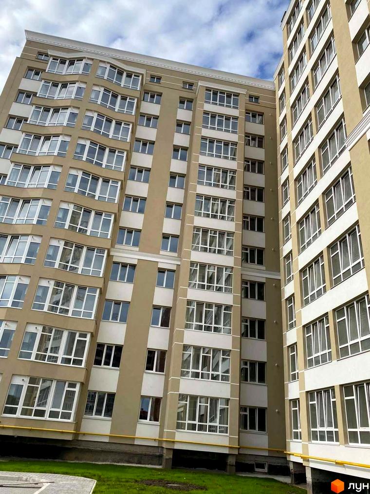 Висотна будівля житлового комплексу з великою кількістю вікон. На території є газон та жовті труби.