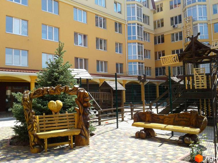 Житловий комплекс Київський маєток: жовтий багатоповерховий будинок з балконами, зелена ялинка та дерев'яні лавки у дворі.