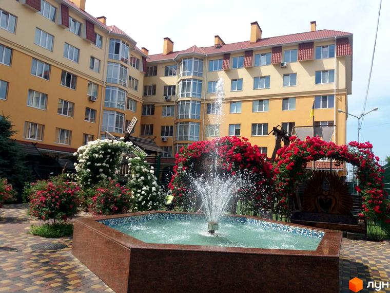 Житловий комплекс Київський маєток з жовтою фасадною частиною та червоним дахом. На прибудинковій території розташований фонтан та арки з червоними трояндами.