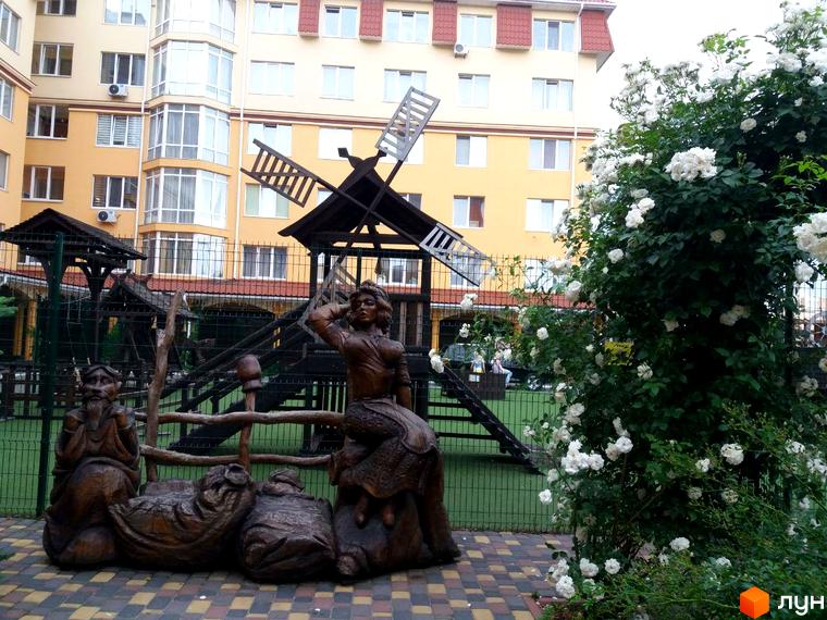 Житловий комплекс Київський маєток з дитячим майданчиком та дерев'яними скульптурами на фоні жовтої багатоповерхівки. Зелені насадження та квіти додають затишку прибудинковій території.