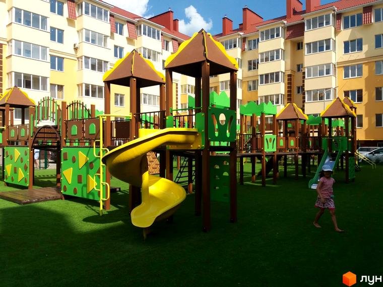 Житловий комплекс Київський маєток: дитячий майданчик з гірками та будинок з жовтою фасадною частиною. Територія навколо новобудови вкрита зеленим газоном.