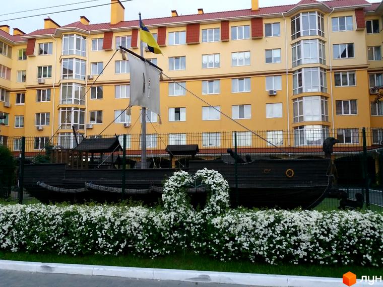 Житловий комплекс Київський маєток з жовтою фасадною стіною, вікнами та декоративним кораблем на прибудинковій території.