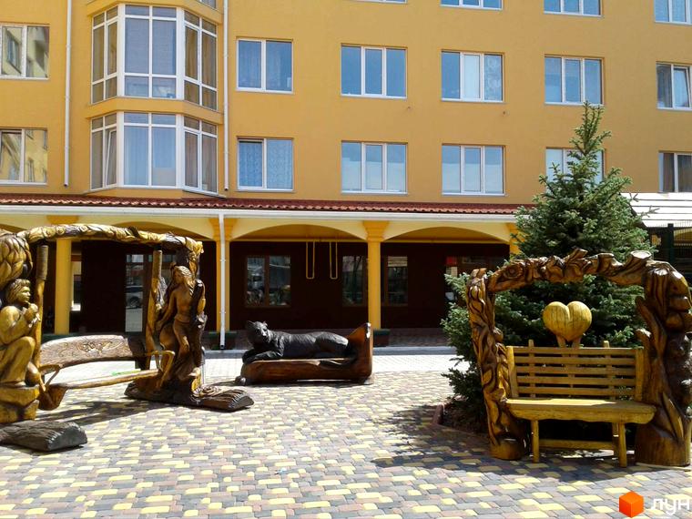 Житловий комплекс Київський маєток: жовта багатоповерхівка з еркерним вікном та зоною відпочинку з дерев'яними скульптурами і лавками.