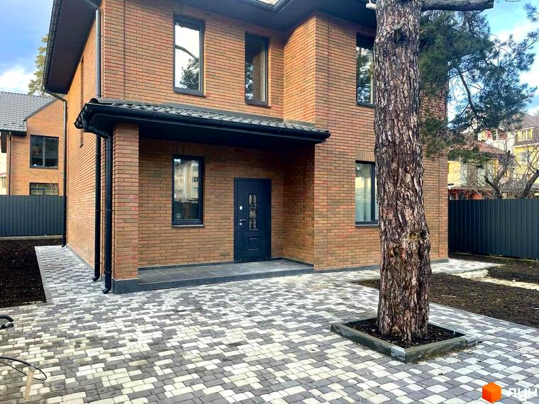 Котеджне містечко КМ Дубовий Гай: двоповерховий будинок з червоної цегли з чорними вікнами та дверима, вимощена доріжка та дерево перед будинком.