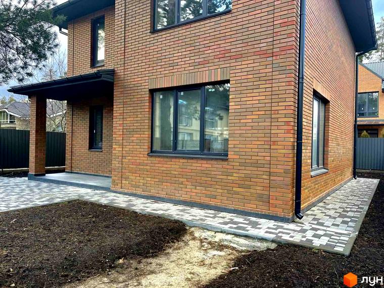 Котеджне містечко КМ Дубовий Гай: двоповерховий будинок з червоної цегли та вимощена прибудинкова територія. Навколо будинку облаштований ландшафт.