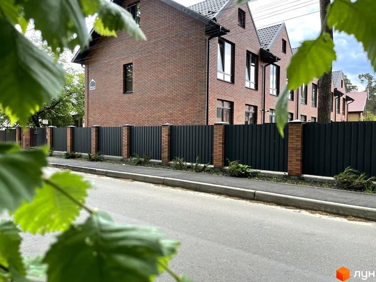 Таунхауси „Новий Brighton“: цегляні фасади та темний металевий паркан з цегляними стовпами. Прибудинкова територія з доглянутою зеленню та асфальтованою дорогою.