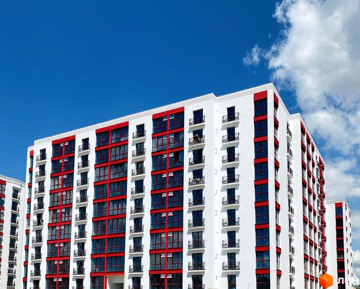 Висотна новобудова з білими фасадами та яскравими червоними акцентами. На балконах встановлені ковані огорожі, а навколо будинку блакитне небо з хмарами.
