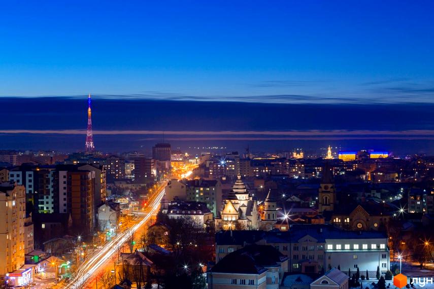 Панорамний вид на місто вночі з телевежею, освітленою в кольори українського прапора. Комплекс Lystopad - це сучасна новобудова, що доповнює міський пейзаж.