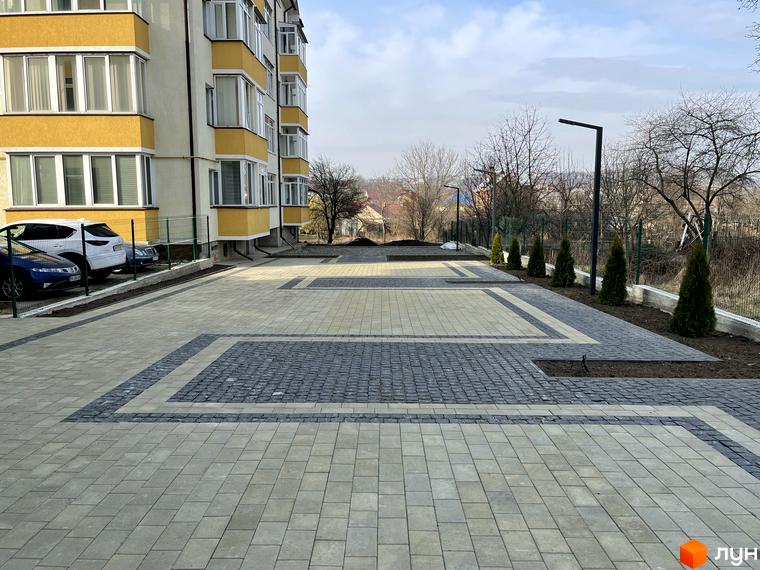 Зовнішній вигляд жовто-білої новобудови ЖК Center з акуратним патіо та паркомісцями. Територія комплексу облаштована бруківкою та зеленими насадженнями.