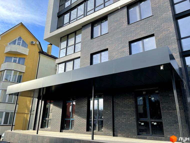 Фасад житлового комплексу Center з сірої цегли та білих панелей, з чорним навісом над входом. Поруч жовта будівля.