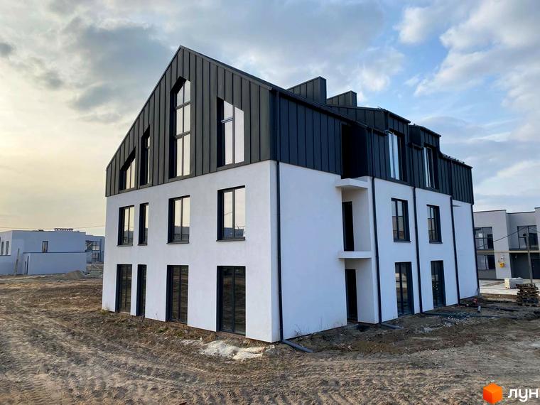 Зовнішній вигляд новобудови ЖК Струмочок 2 з білими стінами та темним дахом, розташованої на будівельному майданчику з ґрунтовою дорогою.