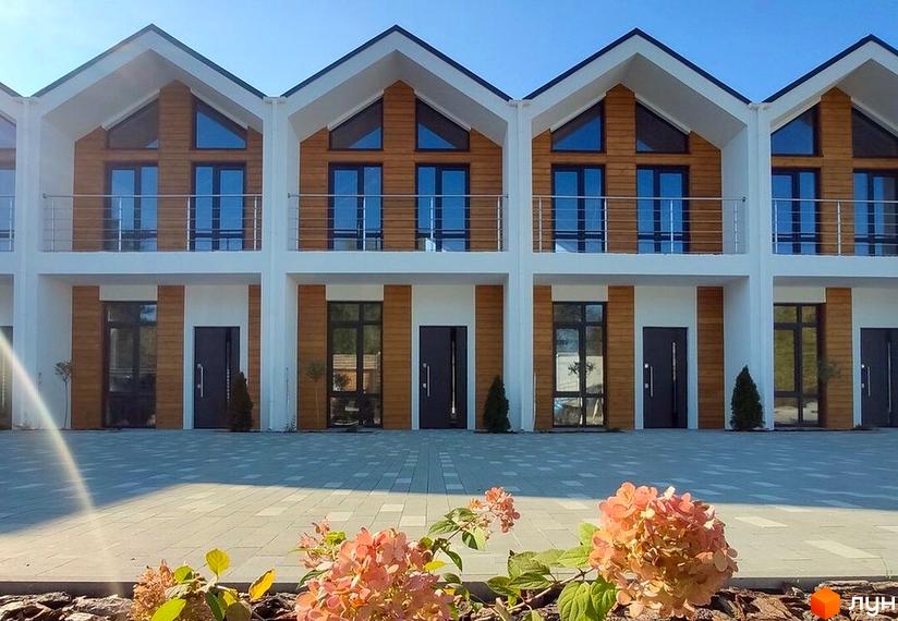 Ряд будинків Котеджного містечка Екохаус з білими фасадами, дерев'яними вставками та балконами. Перед ними брукована територія та квітучі рослини.