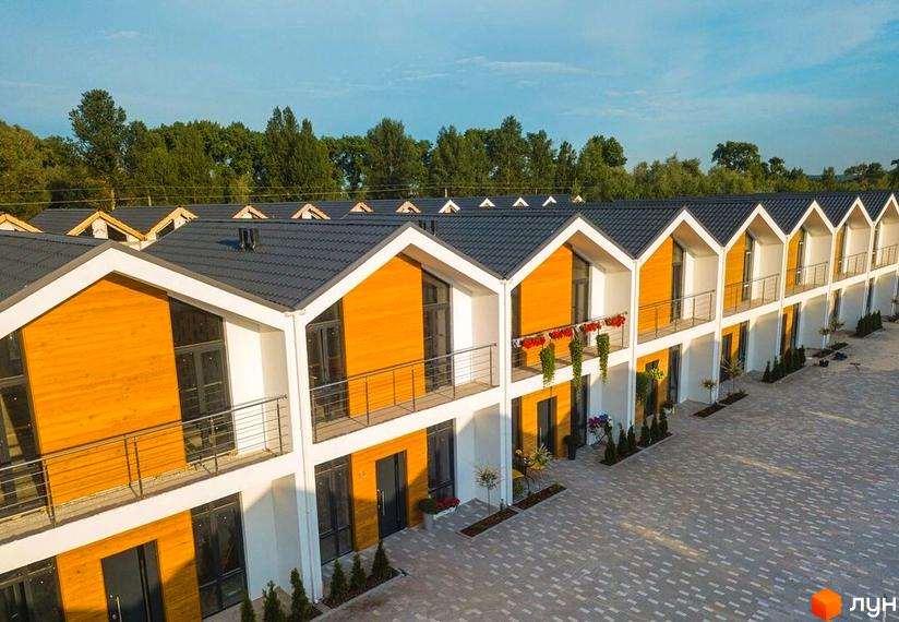 Котеджне містечко Екохаус з рядом будинків, що мають темні дахи, білі фасади та дерев'яні вставки. Перед будинками вимощена територія та зелені насадження.