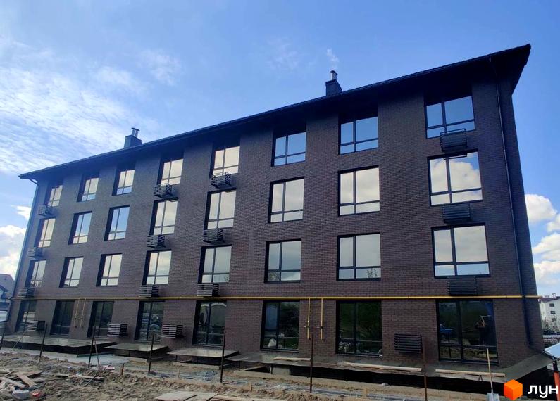 Зовнішній вигляд будинку ЖК Моя Баварія з коричневої цегли та великими вікнами. Навколо будинку видно будівельні роботи та ділянку з піском.