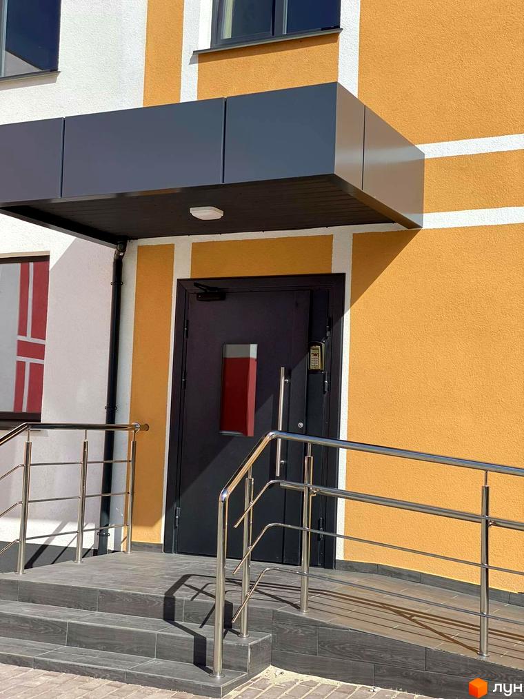 Зовнішній вигляд новобудови ЖК Стайл Хаус з оранжевим фасадом та сірим навісом над входом. Біля входу розташовані сходи з поручнями.