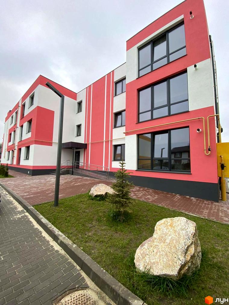 Зовнішній вигляд новобудови ЖК Стайл Хаус з червоно-білим фасадом та великими вікнами. На прибудинковій території зелена галявина з великим каменем.