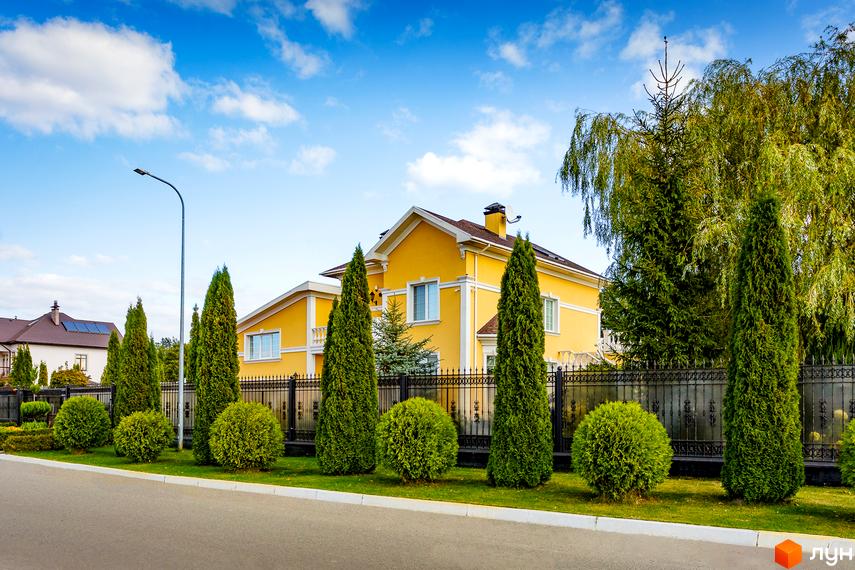 Котеджне містечко КМ Home Resort: жовтий двоповерховий будинок з білими вікнами та дверима, огороджений чорним кованим парканом. Прибудинкова територія доглянута, з газоном та декоративними деревами.