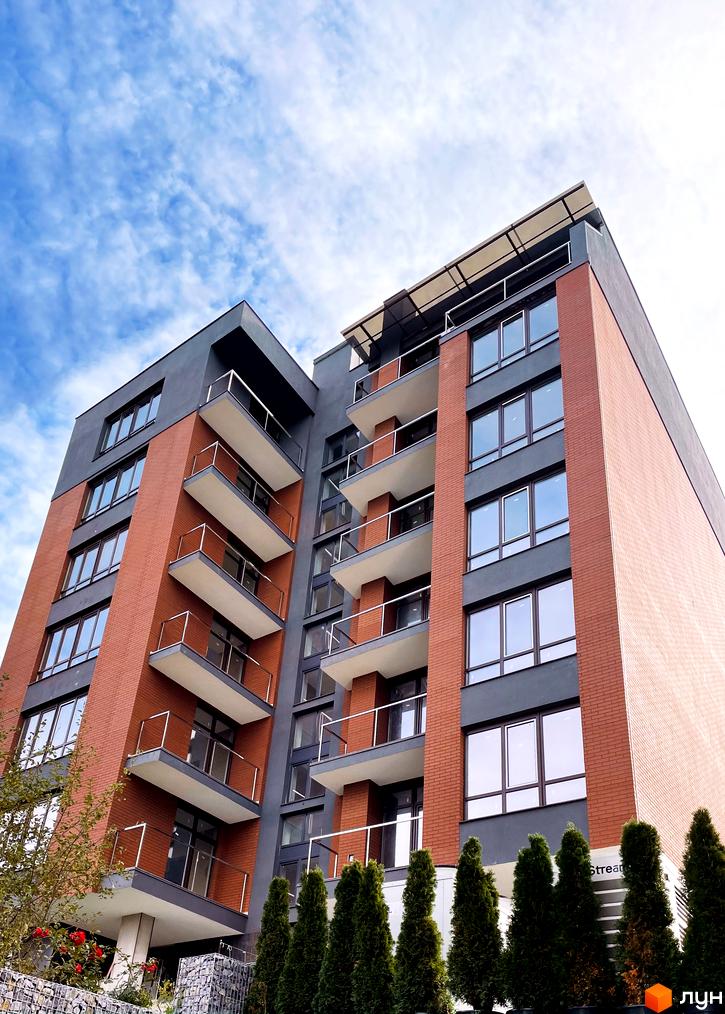 Зовнішній вигляд Central Apartment House з цегляним фасадом та балконами. Поруч висаджені туї та рослини.