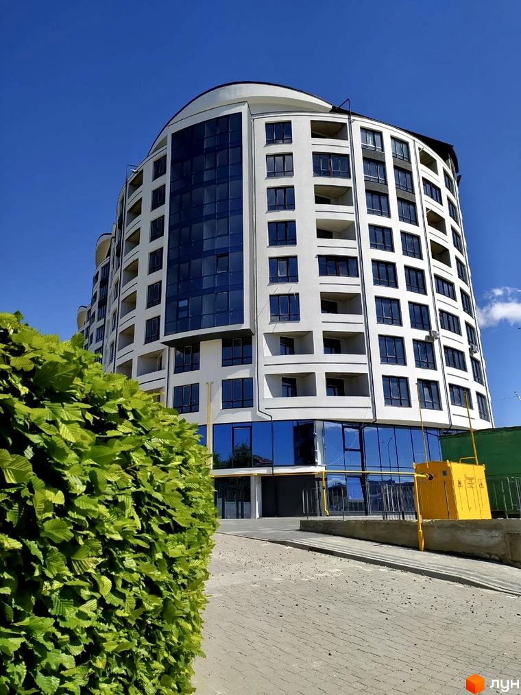 Багатоповерховий житловий комплекс Трускавецька Фортеця з вигнутим фасадом, білими стінами та темними вікнами. Перед будинком зелена огорожа та бруківка.