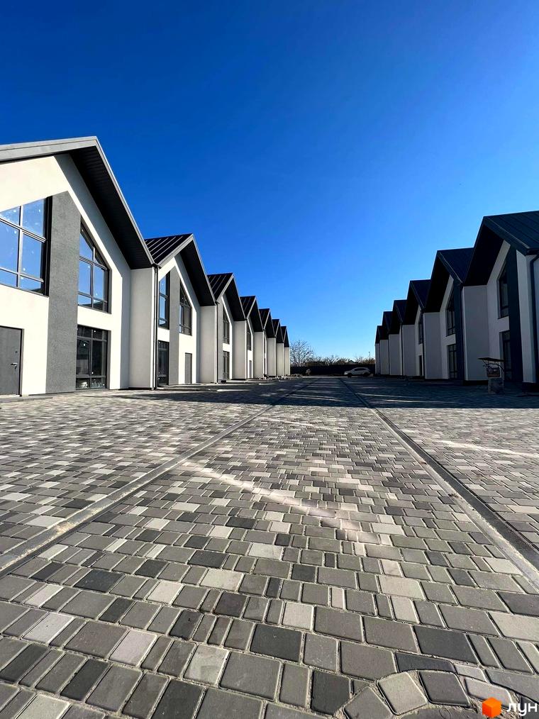 Ряд таунхаусів Scandi Village з білими фасадами, темними дахами та великими вікнами. Перед будинками викладена бруківка сірого та чорного кольорів.