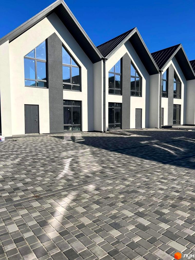 Таунхауси Scandi Village: фасади будинків з білою та темно-сірою обробкою, великими трикутними вікнами та темними дахами. Прибудинкова територія викладена сірою бруківкою.