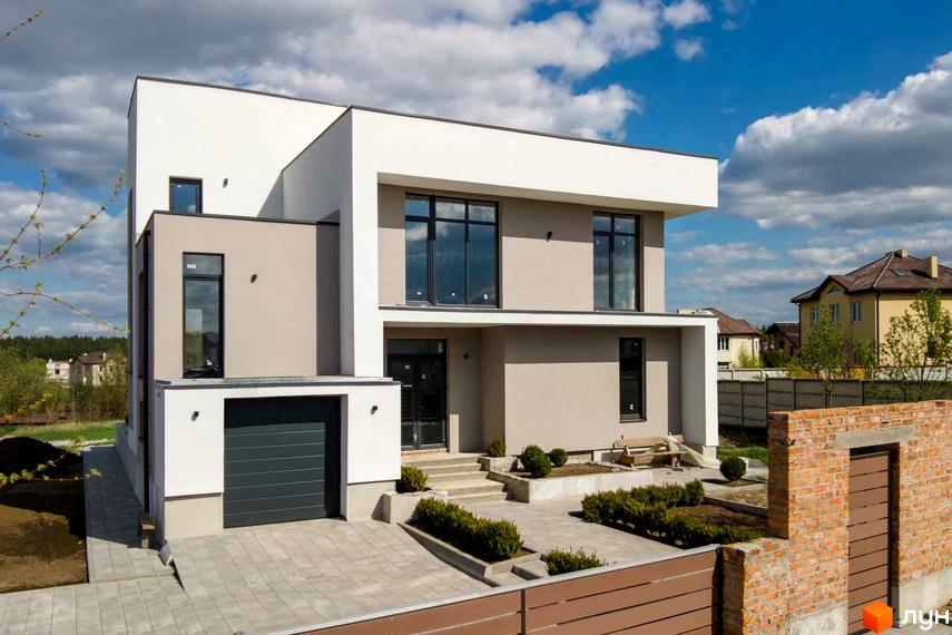 Сучасний двоповерховий будинок з великими вікнами та гаражем. Прибудинкова територія з доглянутими кущами та цегляною огорожею.