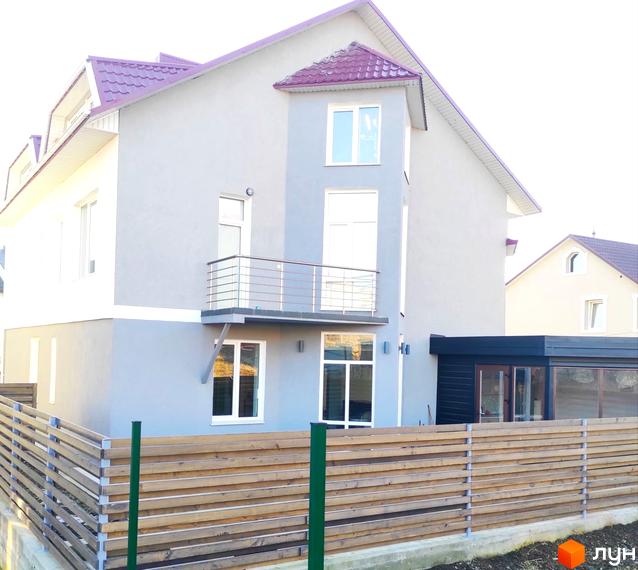 Фасад двоповерхового будинку світло-сірого кольору з бордовим дахом, балконом та великими вікнами. На передньому плані дерев'яний паркан.